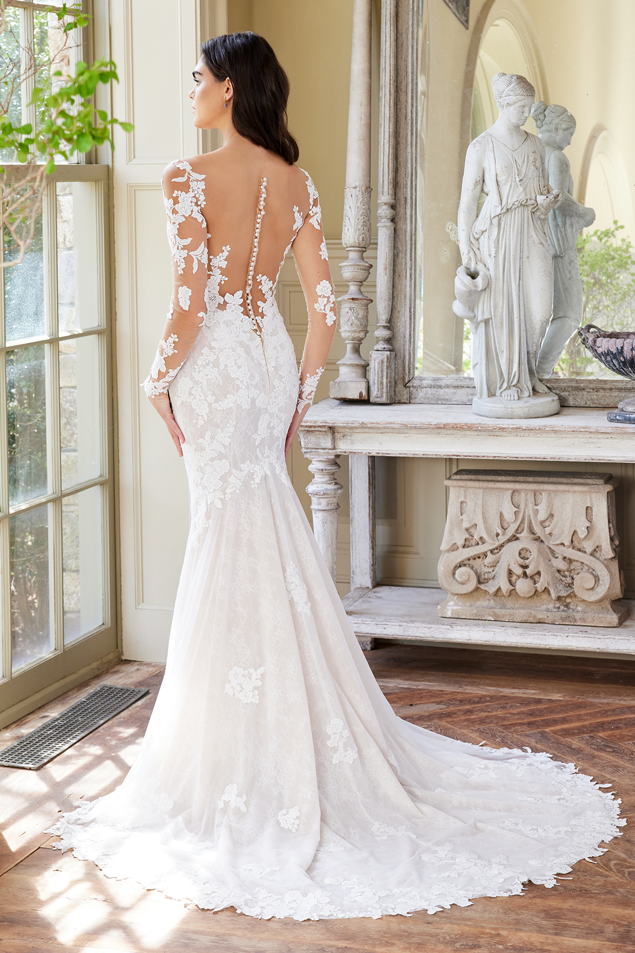 Model is wearing a sleek trumpet wedding gown with long illusion sleeves, a wide v-neckline, and a low illusion back with delicate appliqué detailing.