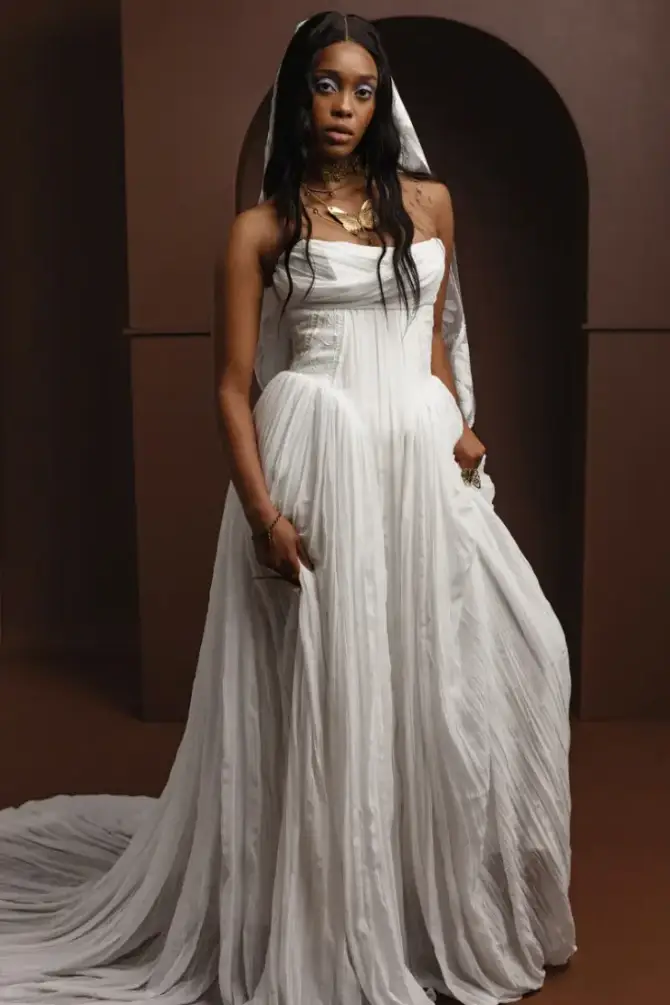 A woman in a flowing white gown and veil stands confidently in front of a dark, arched backdrop. Her expression is serene and regal, emphasizing elegance.