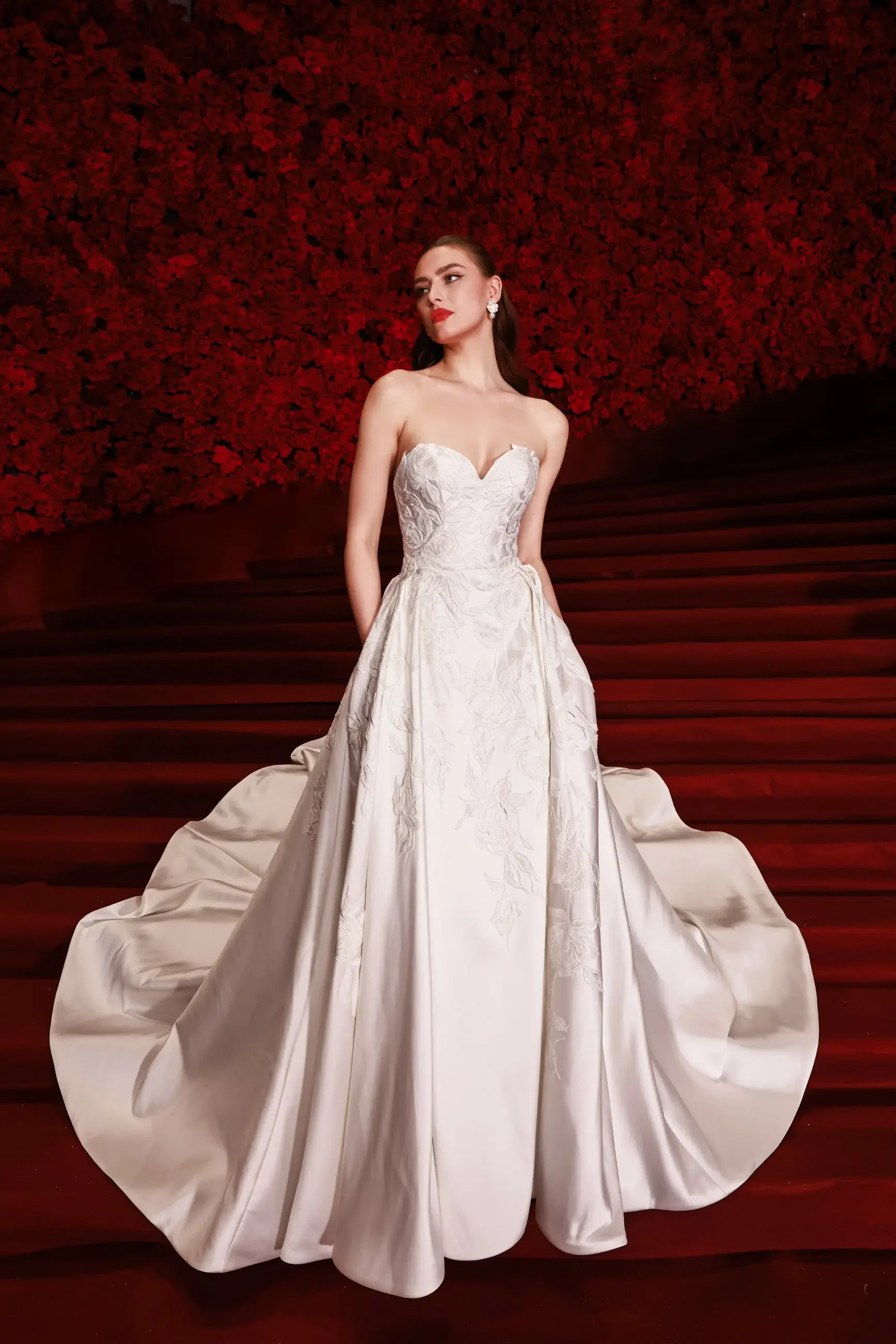 A woman stands elegantly on red-carpeted stairs wearing a strapless, white gown with intricate embroidery. The background is lush with vibrant red flowers.