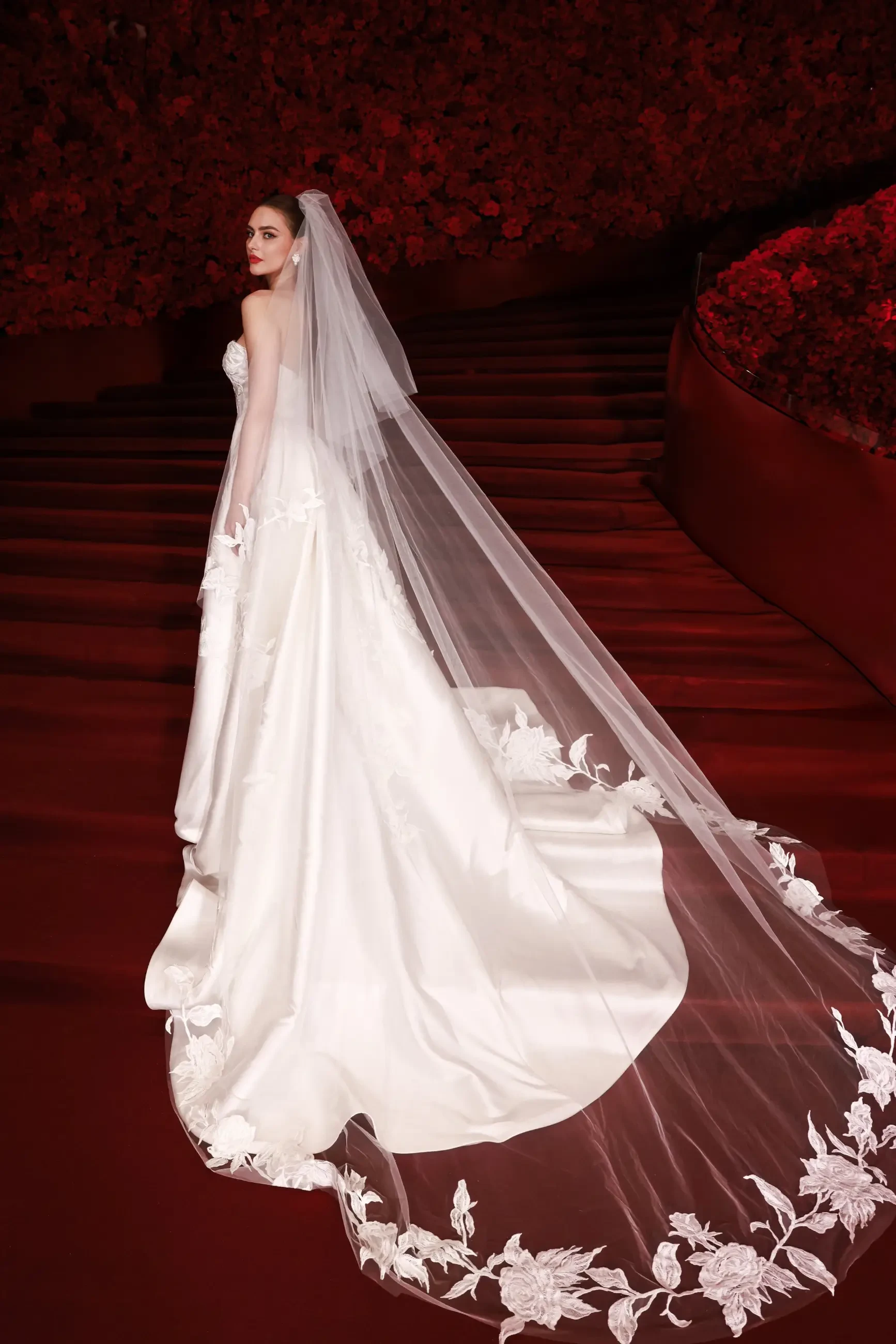 A bride in a flowing white gown with a long, lace-edged veil stands on red-carpeted stairs. The backdrop features abundant red roses, creating a romantic atmosphere.
