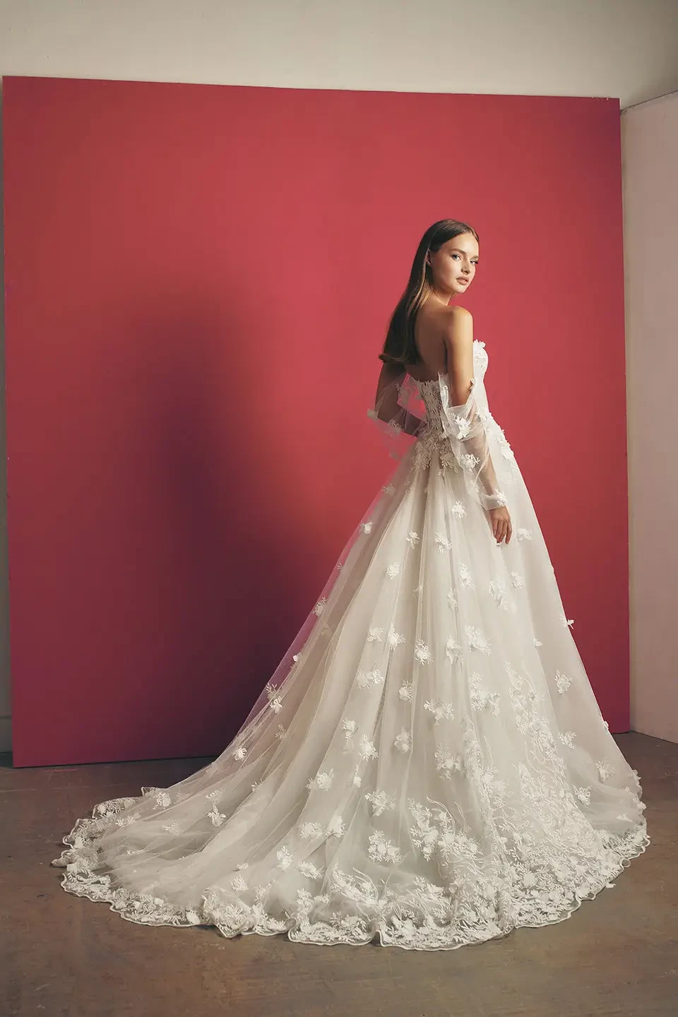 Model is wearing an off-the-shoulder ball gown with a plunging corseted bodice, full lace skirt, sheer puff sleeves, and 3D floral embellishments.