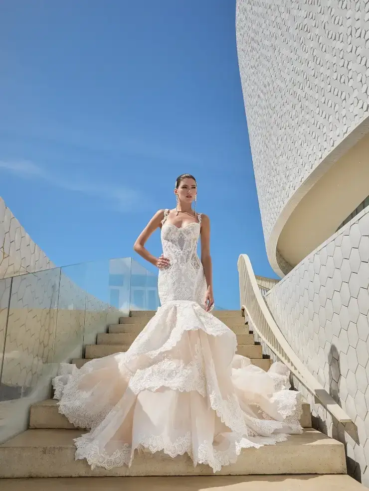 A woman wearing a white wedding dress with intricate lace detailing stands on a staircase against a modern architectural backdrop.