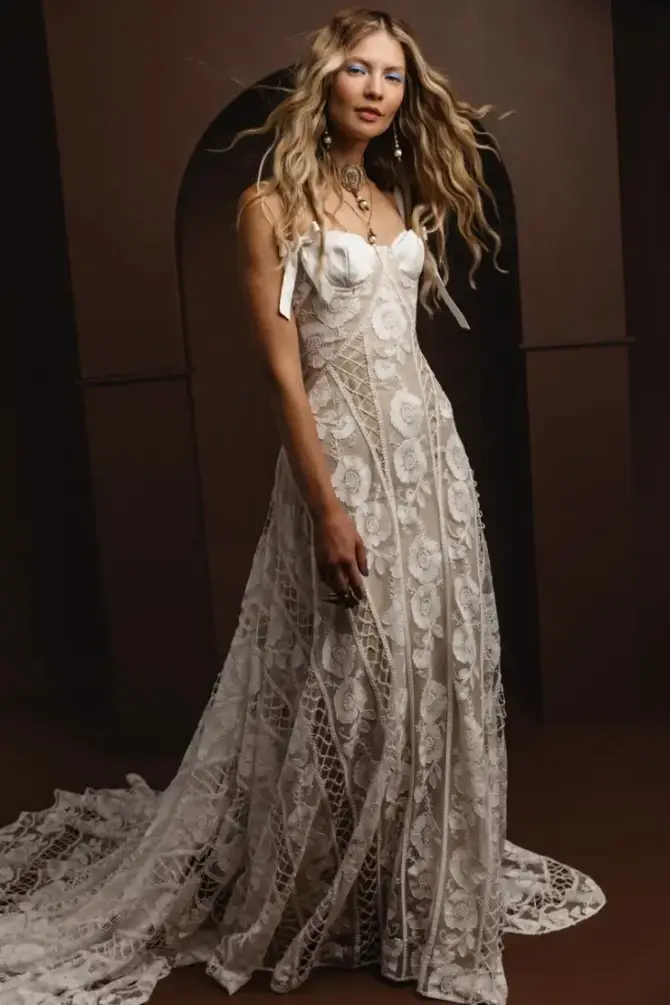 A woman poses in an elegant, flowing lace wedding gown with a long train. Her wavy hair cascades over her shoulders. The background is dark, highlighting the intricate dress details.
