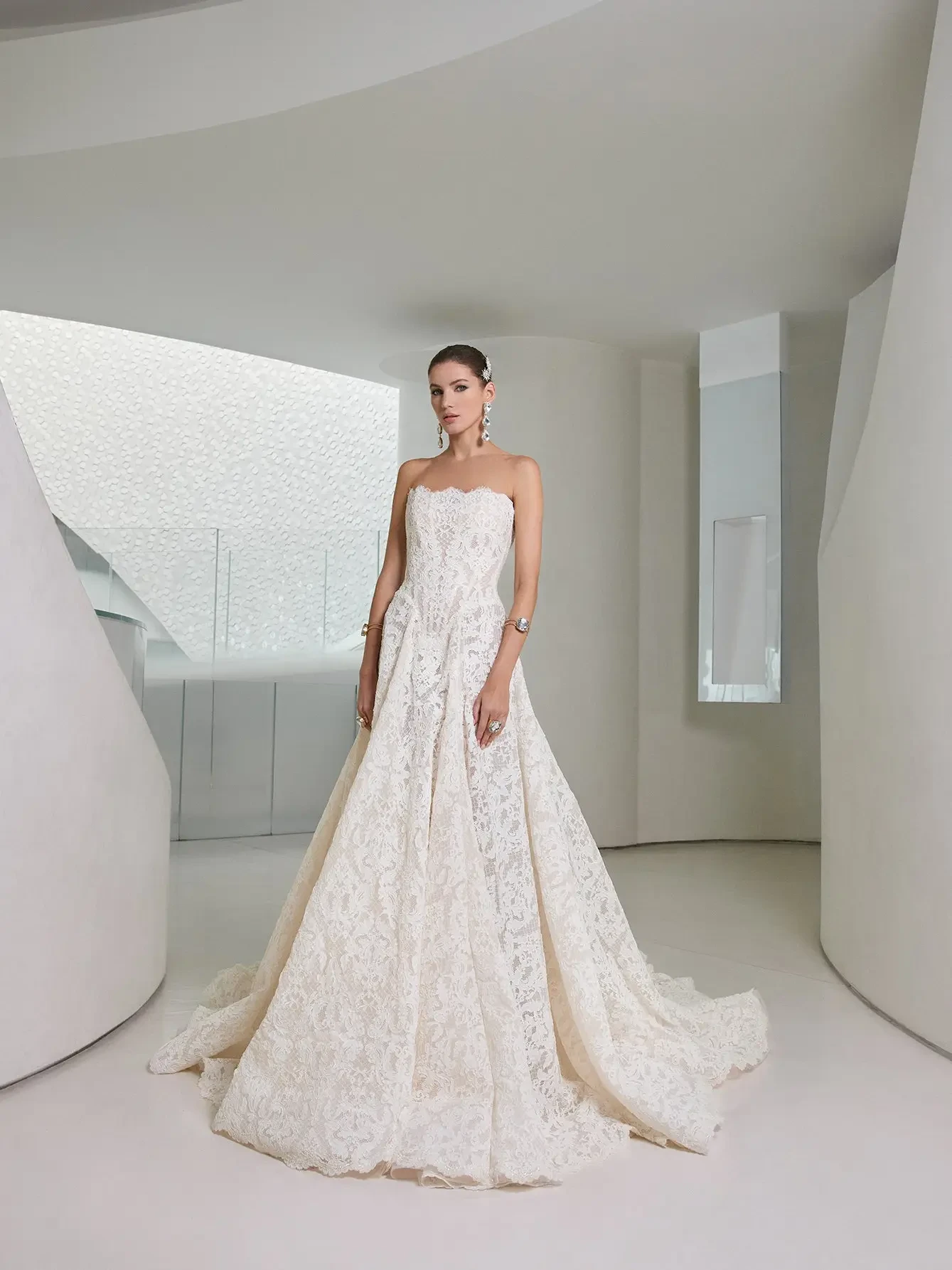 A woman in a strapless lace wedding gown stands in a sleek, modern interior with white curved walls, exuding elegance and sophistication.