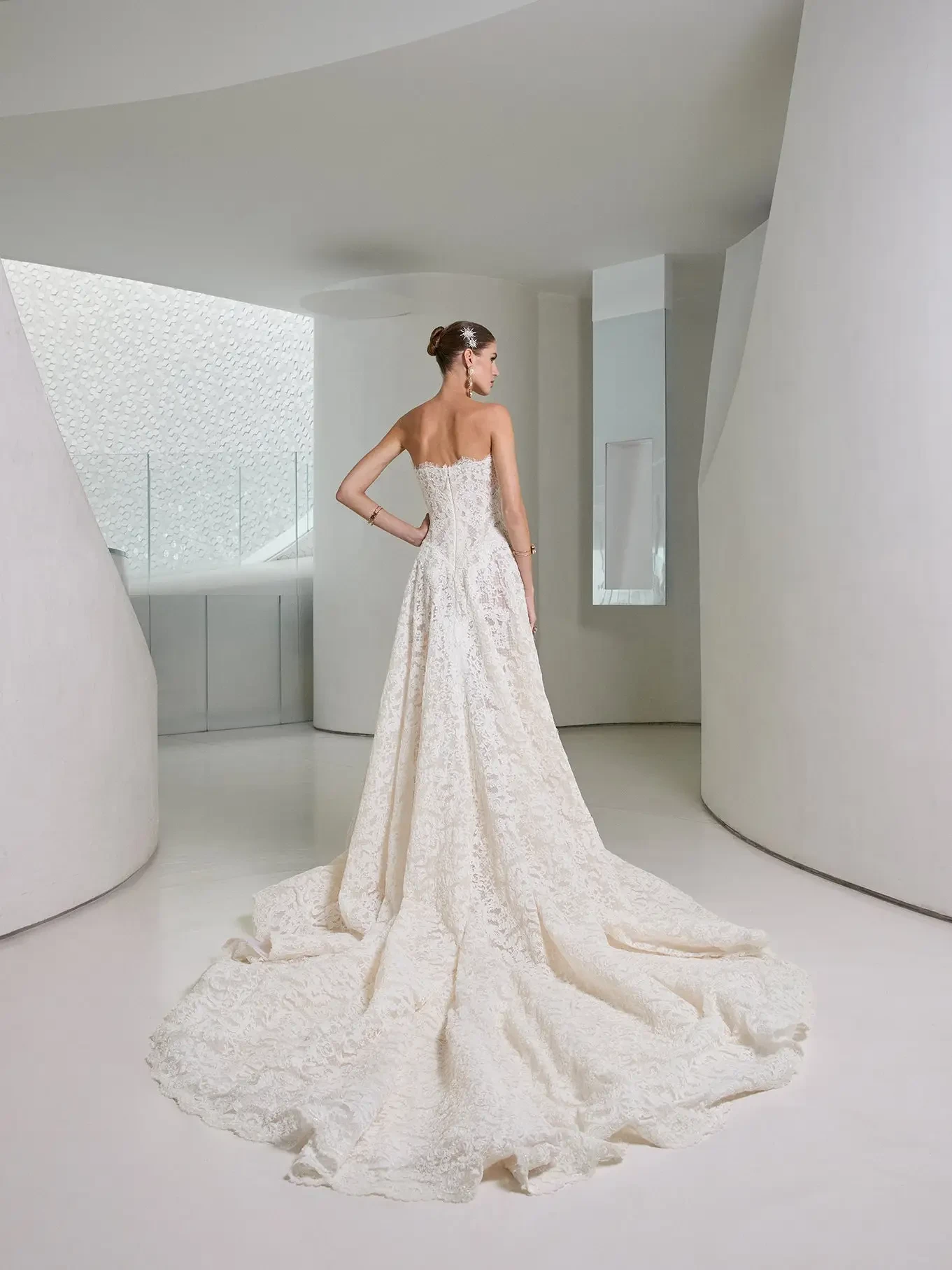 A bride in a luxurious, strapless lace gown with a long train stands gracefully in a modern, curved hallway, exuding elegance and serenity.