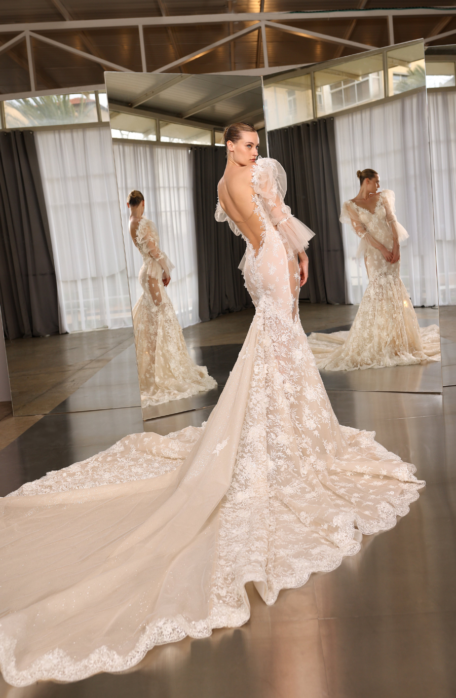 Model is wearing an off-the-shoulder mermaid gown with an illusion corset back, decorated sleeves, and a tulle train.