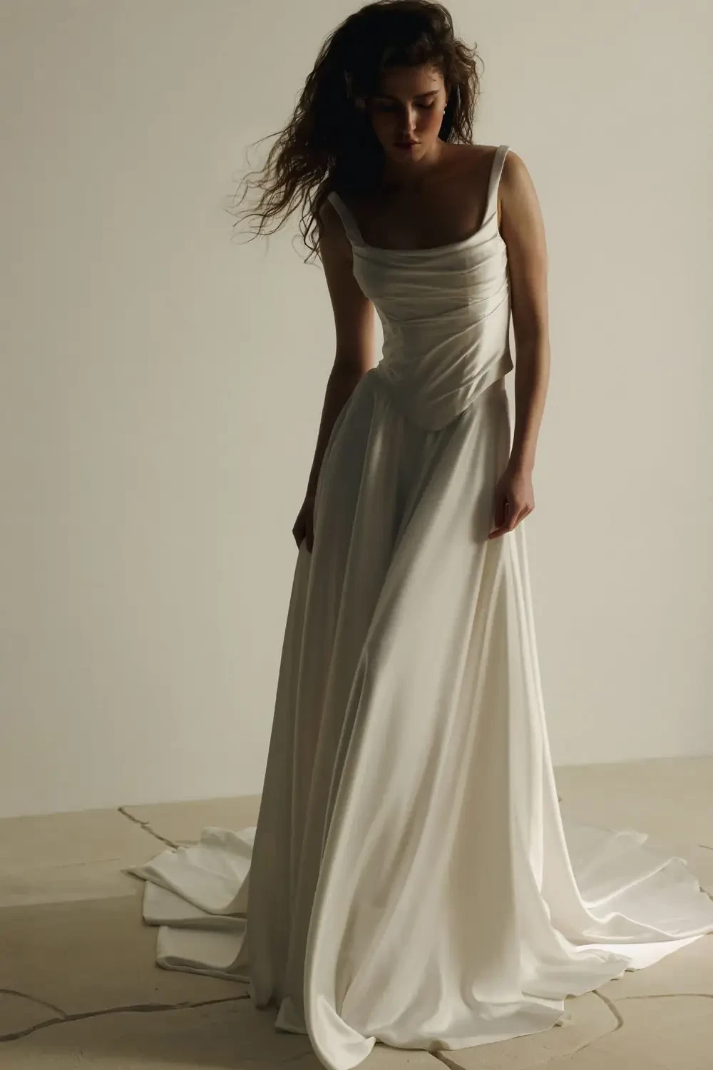 A woman in an elegant white gown stands on a light stone floor, gazing downward. Her relaxed pose and soft lighting create a serene, graceful atmosphere.
