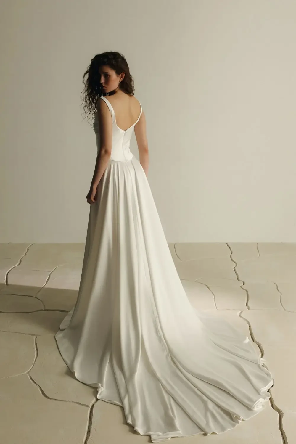 A woman with curly hair wears a flowing white gown, turned slightly away on a textured, cracked floor. The setting feels elegant and serene.