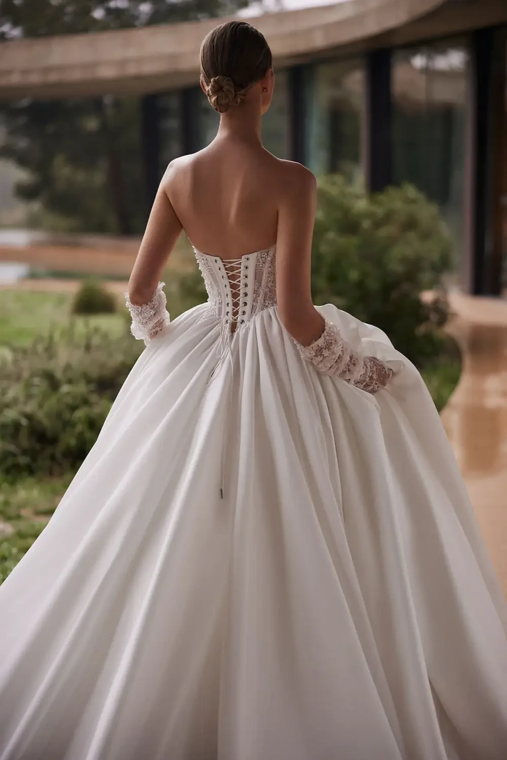 A woman in a strapless, flowing white wedding gown with lace-up back stands outdoors. Her hair is in an elegant bun, conveying a romantic and serene tone.
