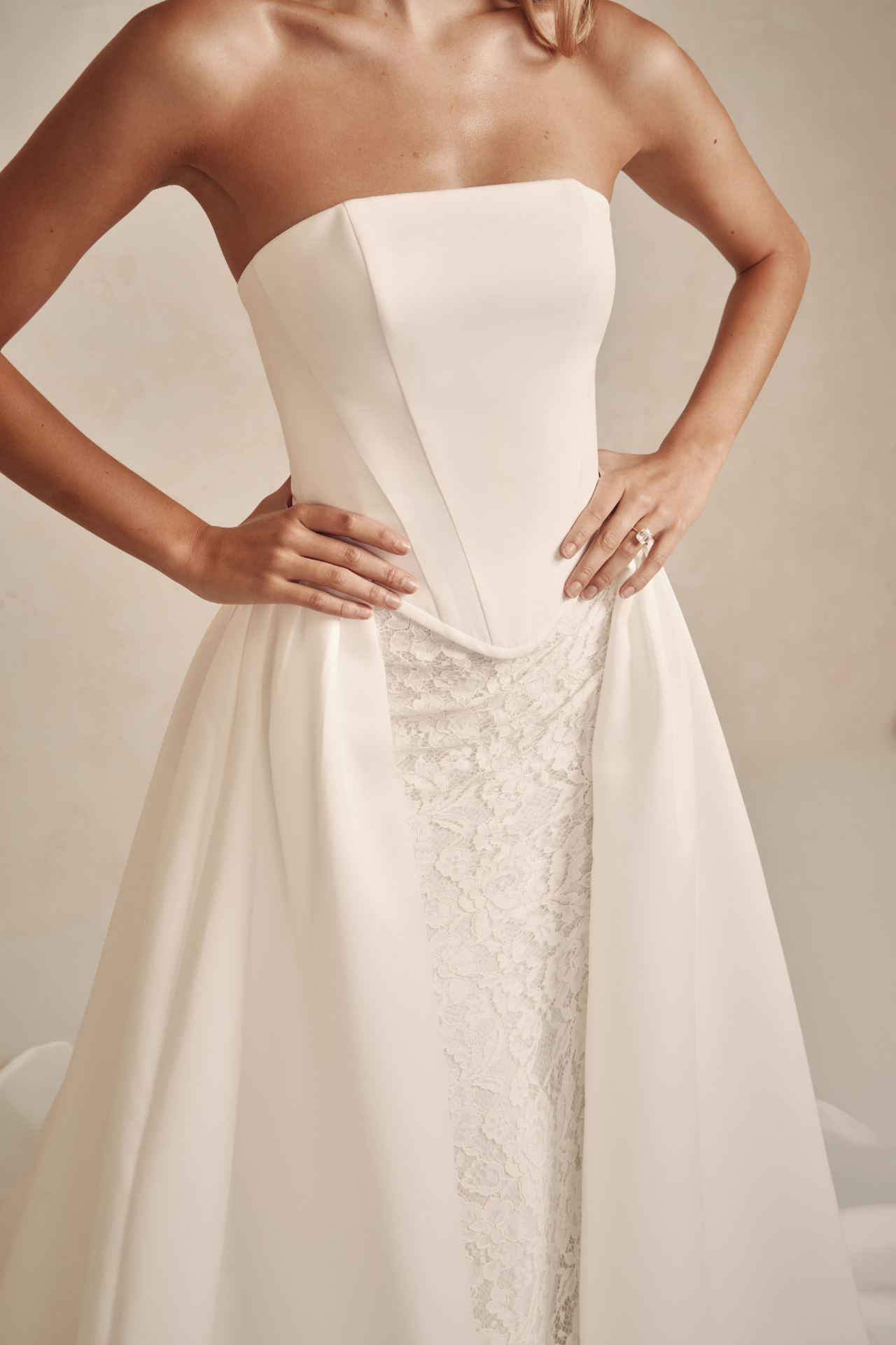 Model is wearing a strapless duchess satin gown with a sculpted basque waist, fitted lace underlayer, and detachable overskirt.