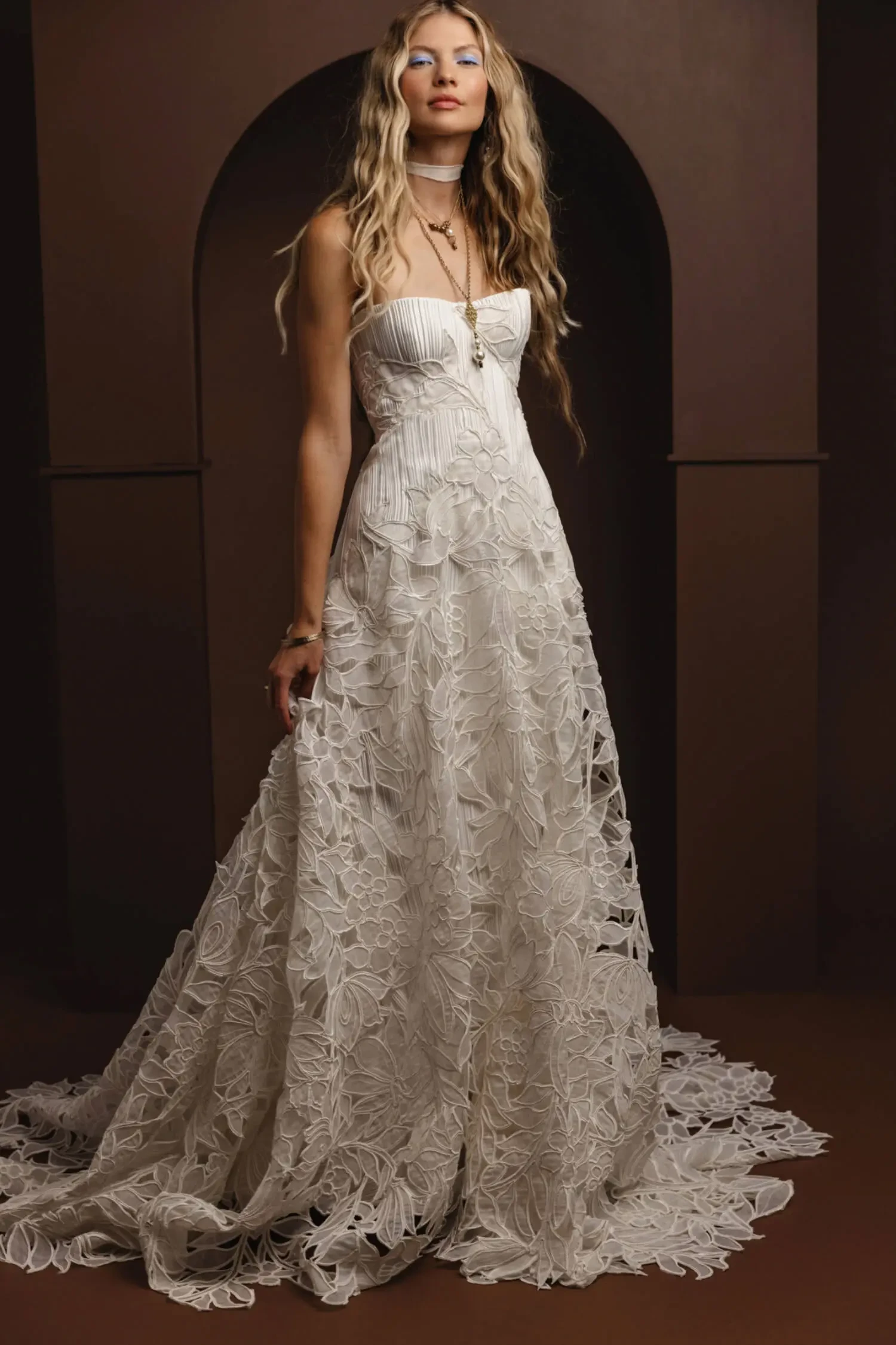 A woman in a textured, strapless white gown with floral patterns stands confidently in an arched, brown backdrop. Her long hair is styled in loose waves.