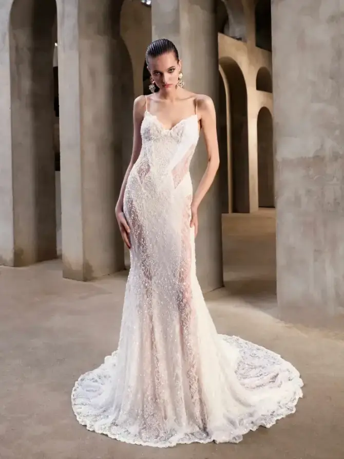 A woman stands in a stunning wedding dress with intricate lace details, showcasing a fitted silhouette and a train, set in an elegant architectural space with arches and neutral tones.