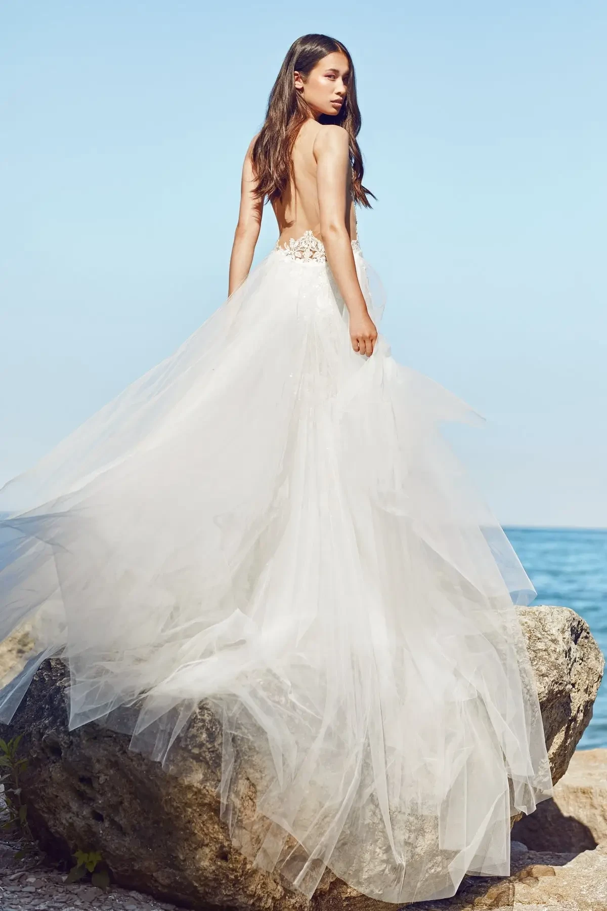 Model is wearing a trumpet-style wedding gown with an illusion lace back, fitted silhouette, and a detachable overskirt