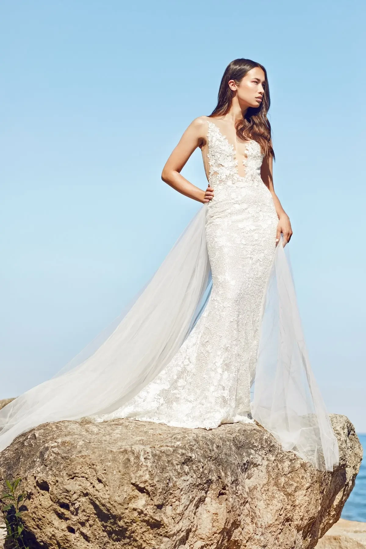 Model is wearing a trumpet-style wedding gown with an illusion lace back, fitted silhouette, and a detachable overskirt